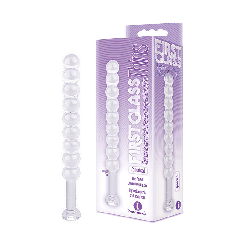 A transparent glass massager, named "The 9's First Glass Thins, Spherical - Clear Glass 17.8 cm Anal Beads," is displayed next to its packaging. The predominantly purple and white packaging features the text "FIRST GLASS" and highlights its spherical design. The toy consists of a string of connected spheres that gradually increase in size, making it ideal for temperature play.