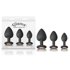 Three black silicone plugs of varying sizes are lined up beside packaging labeled "Glams Spades Butt Plug Trainer Kit." Each bejeweled silicone plug base is adorned with a colorful, gem-like decoration. The three-piece set and its plugs are displayed on a plain white background.