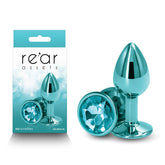A packaged set of lightweight aluminum, teal anal toys with a gemstone finish on the base. The packaging displays the brand name "Rear Assets - Small - Teal" and showcases the body-safe product. The plugs are shown both inside the packaging and separately on the right side.