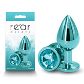 A teal-colored, gem-encrusted, lightweight anal toy labeled "Rear Assets - Medium - Teal" is displayed next to its packaging, which showcases the product's image and the branding "Rear Assets" by NS Novelties. Both the toy and its packaging feature a matching shiny, turquoise blue aesthetic.