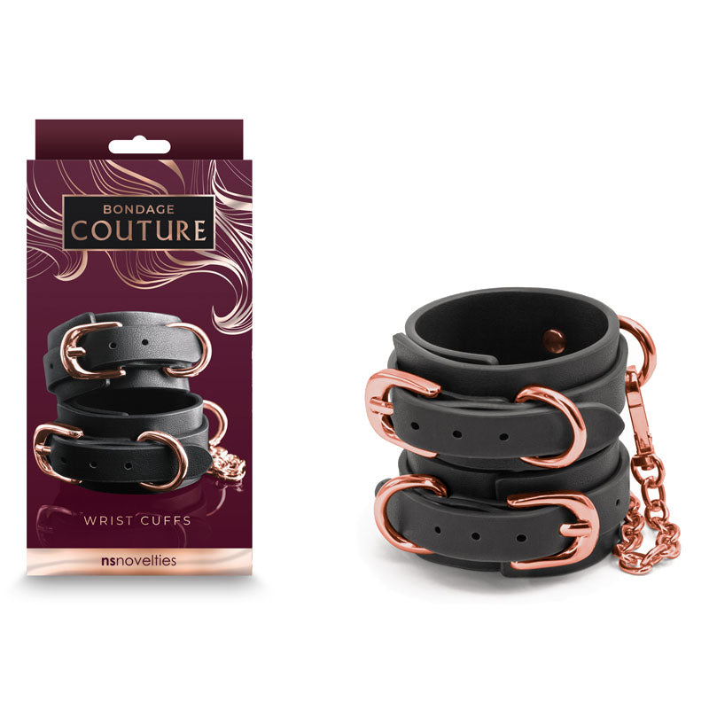 A pair of Bondage Couture Wrist Cuffs - Black, crafted from synthetic material with rose gold metal buckles and a connecting chain, displayed next to the dark purple packaging. The packaging prominently features the text "Bondage Couture Wrist Cuffs" along with "NS Novelties" at the bottom.