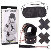 Image of the "Sex & Mischief Brat Kit" from Sportsheets, featuring wrist restraints, an eye mask, two black pasties, and a small mini flogger. The packaging includes an image of a person wearing the items on the front.