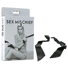 A product box labeled "Sex & Mischief Silky Sashes Black" showcases a black-and-white image of a person holding luxurious silk sashes. Beside the box, there is a detached, long, black silky ribbon. The text on the box reads "Silky Sashes Black.