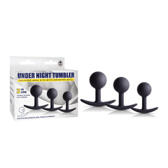 A box of "Under Night Tumbler Butt Plug Set" is displayed alongside its contents: three black silicone anal plugs of different sizes, each featuring a weighted metal ball at the top and an ergonomic base. The box highlights product details and emphasizes a "3 in 1" feature.