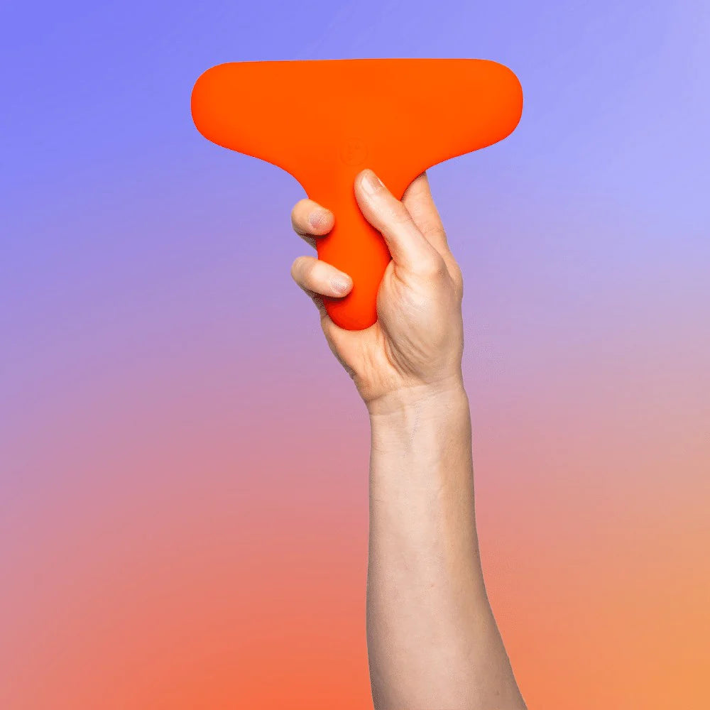 A hand holds the Ziggy™ vibrator, a vibrant orange T-shaped object made of medical-grade silicone, against a gradient background of blue and orange hues. The Ziggy™ has a wide, rounded top and a narrower bottom section, being held securely in an upright position.