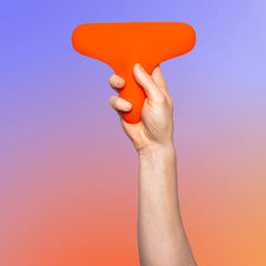 A hand holds the Ziggy™ vibrator, a vibrant orange T-shaped object made of medical-grade silicone, against a gradient background of blue and orange hues. The Ziggy™ has a wide, rounded top and a narrower bottom section, being held securely in an upright position.