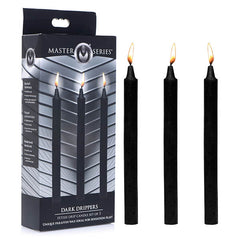 A box of Master Series Fetish Drip Candles Black Sex Wax is shown with three tall, black candles standing upright next to it. Made with a paraffin wax formula, the candles have yellow flames, and the packaging is predominantly black with a grey and silver pattern—perfect for sensation play.
