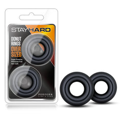 A product package labeled "Stay Hard - Donut Rings Oversized" containing two black circular rings is displayed. Next to the package are two additional black rings positioned upright. The packaging highlights super stretchy, phthalate-free, skin-safe materials designed for achieving harder erections that last longer!