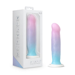 The Avant - D17 - Lucky, a pastel-colored silicone dildo in shades of pink, blue, and purple, is showcased inside white packaging with the brand name "Avant Lucky" at the bottom. The packaging features a clear window to view the product, includes icons for various specifications, and indicates that it is harness compatible.