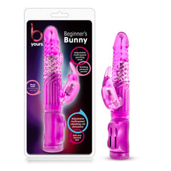A packaging containing a pink, rabbit-shaped vibrator labeled "B Yours - Beginner's Bunny Rabbit Vibrator" is displayed. This transparent rabbit vibe features a curved tip for G-spot stimulation and clit-stimulating rabbit ears. The packaging highlights its multi-speed settings and soft, flexible texture.