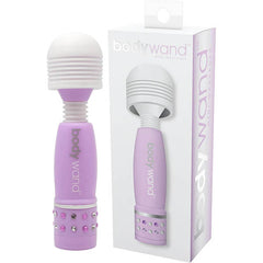 The image shows the Bodywand Mini - Lavender Mini Massager Vibrator, featuring a purple body, soft-touch white head, and gem accents at the base, positioned next to its packaging. The packaging displays an image of the massager with "body wand" written in light purple text. This device offers multi-directional usage for strong stimulation.