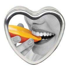 Heart-shaped metal tin featuring a black-and-white image of a person’s smiling mouth about to bite into a vibrant orange mango slice. The hand holding the mango has nails painted in a light color. Inside, you'll find the Edible Mango Massage Candle, infused with hemp seed oil for added relaxation and perfect for vegan massage experiences.