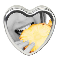 A heart-shaped metal tin, bearing an image of someone about to bite into a vibrant pineapple slice against a black-and-white backdrop, highlights the refreshing appeal of the Edible Pineapple Massage Candle.