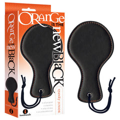 The Orange Is The New Black - Spanky Junior, a black leather paddle with an ergonomic handle and smooth paddle surface, is displayed beside its packaging. The packaging showcases bold text that reads "Orange is the New Black" in a mix of white and black letters on an orange background. The paddle features a stitched edge and includes a small cutout for hanging.