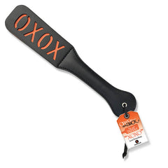 The 9's Orange Is The New Black, Slap Paddle XOXO features a black PU leather paddle with the word "XOXO" reverse cut in red letters. The handle comes attached to a hanging tag with orange and white design and text that details product branding and information.