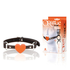 A heart-shaped orange silicone gag attached to an adjustable black strap is displayed alongside its packaging. The package highlights the product's name, "The 9's Orange Is The New Black, Heart Gag," and includes a graphic showing the gag in use—ideal for those new to beginner gag-play.
