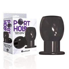A black cylindrical object with a rounded top and flat base is placed in front of its packaging. The packaging reads "The 9's Port Hole Hollow Butt Plug - Black" and highlights "Peeker Plugs in Body-Safe TPR." Designed for adult use, this smooth hollow tunnel plug is ideal for lube insertion, with the box showcasing it from different perspectives.