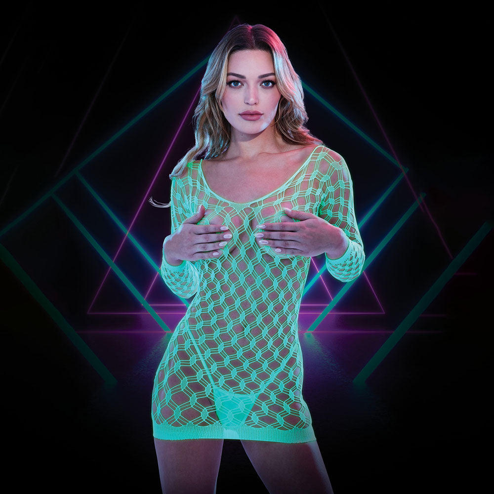 A woman stands confidently in front of a geometric neon background. She is wearing a LAPDANCE Glow In The Dark Mini Dress - 1Size, featuring a bright green fishnet-style design that covers her upper body and thighs. She has long hair and a focused expression, set against the neon backdrop adorned with intersecting lines and triangles.