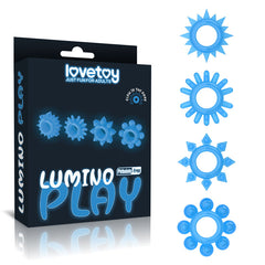 A black box labeled "Lumino Play Penis Rings 4 Pack" features four blue glowing rings with various spike patterns. The text on the box reads "Lovetoy - Just Fun For Adults" and "Phthalate-Free" in white. Each glow-in-the-dark penis ring's unique design is displayed to the right of the box.