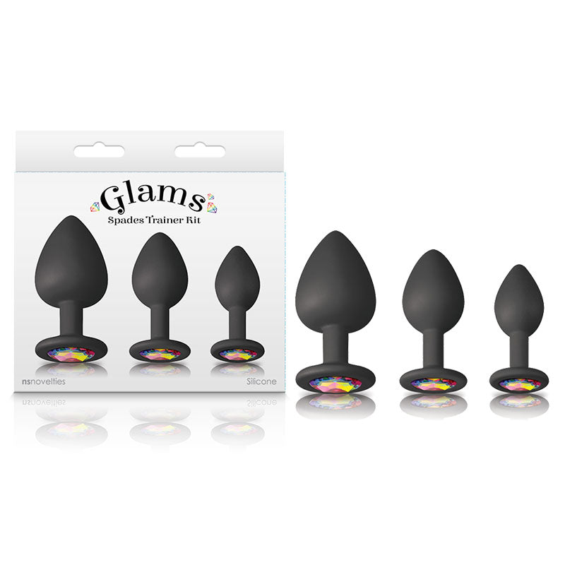 Three black silicone plugs of varying sizes are lined up beside packaging labeled "Glams Spades Butt Plug Trainer Kit." Each bejeweled silicone plug base is adorned with a colorful, gem-like decoration. The three-piece set and its plugs are displayed on a plain white background.