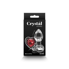 A product image showcasing a black box labeled "Crystál DESIRES" with the words “nü Sensuelle” and “GLASS” at the bottom. The package displays a 7.3 cm clear glass butt plug adorned with a red heart-shaped gem at its base, perfect for an intimate experience. The gem is prominently featured beside the plug.