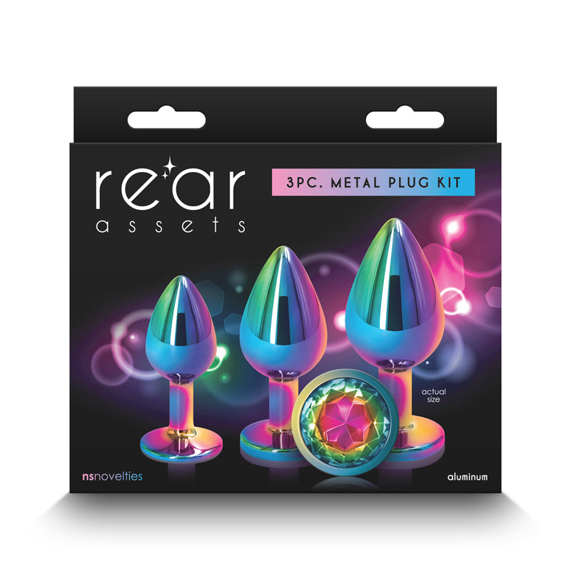 Packaging for the "Rear Assets Butt Plug Trainer Kit - Multicolour - Rainbow" by nsnovelties. The box showcases three iridescent, body-safe butt plugs adorned with gem-like features against a dark backdrop enhanced by vibrant light effects. The text highlights "actual size" and the brand name, "nsnovelties".