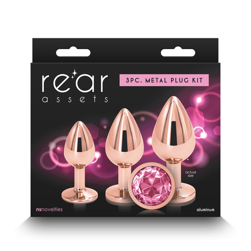 An image showcases the Rear Assets Trainer Butt Plug Kit in rose gold with pink, consisting of a 3-piece metal plug set. The chrome-plated plugs have a glossy rose gold finish and feature pink jewel-like ends. The packaging presents the product against a black background adorned with decorative pink lights, and highlights compatibility with "nsnovelties" and "aluminum," confirming that they are body-safe.