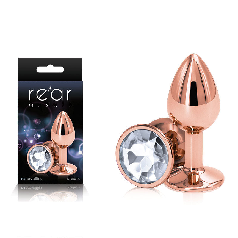 The Rear Assets Rose Gold Small Butt Plug, a metallic anal toy with a jewel-like decoration at its base, is displayed next to its packaging. The lightweight and body-safe product's packaging features an image of the butt plug along with the text "rear assets" and "nsnovelties.