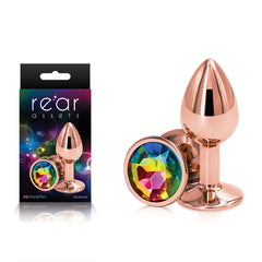 The Rear Assets Rose Gold Small Butt Plug features a gem-like multicolored jewel at its base. The product packaging showcases the same item, prominently displaying the brand names "Rear Assets" and "ns novelties." This lightweight, chrome-plated plug is crafted from aluminum.