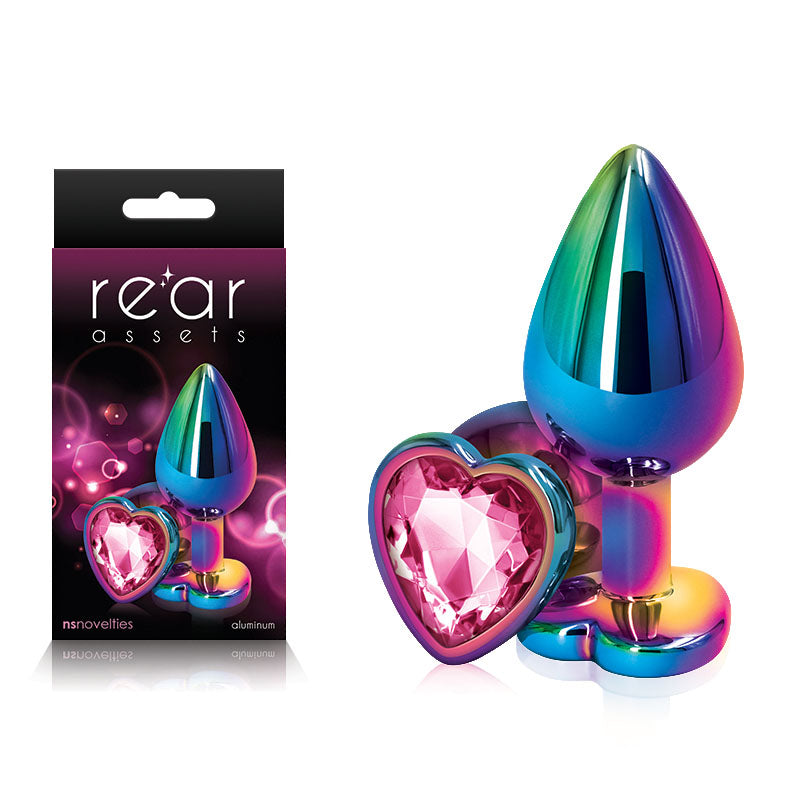 A metallic rainbow-colored butt plug, named the Rear Assets Multi Coloured Heart Butt Plug, features a pink heart-shaped gem and is pictured next to its box. The box displays the product image along with the text "rear assets" and "nsnovelties" against a black and purple background. This body-safe anal toy is crafted from high-quality aluminum for a luxurious experience.