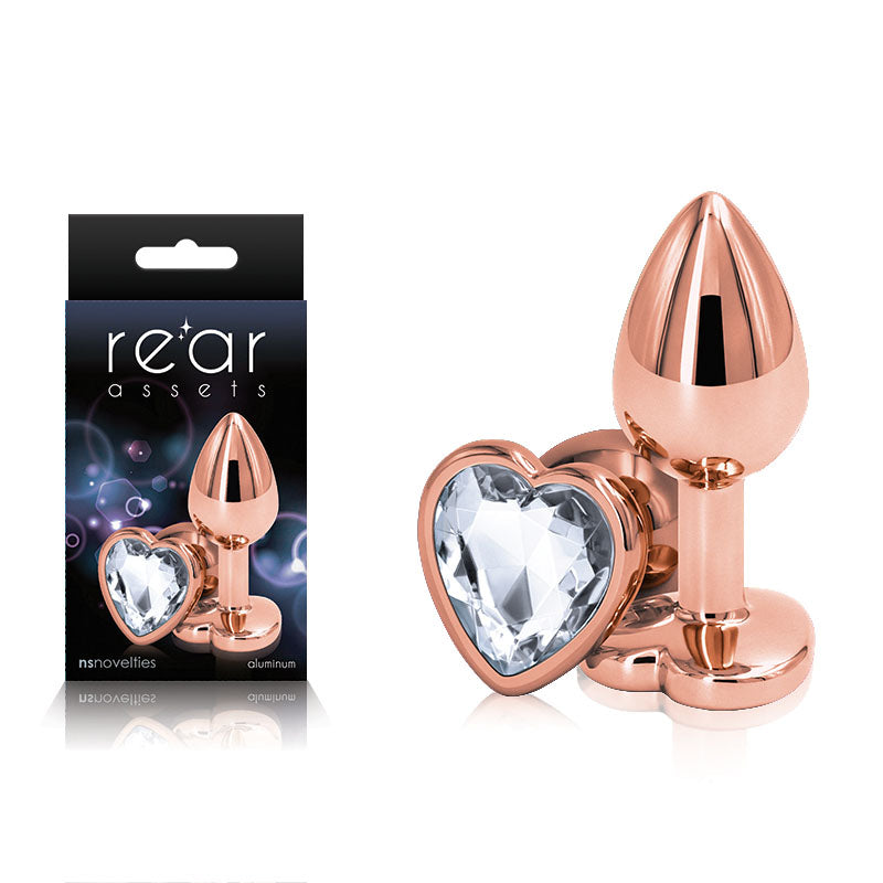 A product image featuring the Rear Assets Rose Gold Heart Small Butt Plug, made of lightweight aluminum in a metallic rose-gold finish with a heart-shaped clear gem at the base, alongside its packaging labeled by NS Novelties. The body-safe packaging, showcasing the same item, has a dark background with reflective highlights.