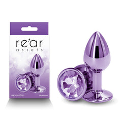 A product photo of the "Rear Assets - Small - Purple," an aluminum butt plug featuring a clear gemstone at its base, displayed both in real form and on its packaging. The packaging showcases the brand name "Rear Assets" and emphasizes that "NS Novelties lightweight aluminum anal toys" are crafted from body-safe materials.