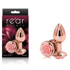 A shiny, rose gold "Rear Assets Rose - Small - Rose Gold 7.6 cm Metal Butt Plug" adorned with a pink rose at the base is shown. The lightweight aluminum item is displayed both outside and partially inside its black and pink packaging, which bears the brand name "Rear Assets" along with mentions of "nsnovelties" and "body safe anal toys".