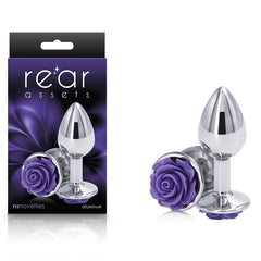 The image shows a chrome 7.6 cm metal butt plug with a purple rose decoration at the base. This small, body-safe butt plug is presented in its black and purple packaging, labeled "Rear Assets," as well as separately outside the box. This elegant piece of anal toy craftsmanship is made by NS Novelties.