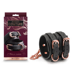 A box of Bondage Couture Ankle Cuffs - Black by NS Novelties is displayed. The cuffs, crafted from sleek black synthetic material, feature rose gold buckles and an accompanying chain. The elegant dark packaging showcases an image of the cuffs with the labels "Bondage Couture" and "Ankle Cuffs.