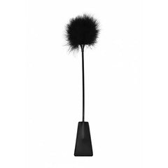 The Ouch! Crop With Feather is a black whip featuring a leather handle, with a fluffy, fur-like tuft at one end and a wide, flat paddle at the other. Photographed against a plain white background, its sleek and distinctive design is showcased beautifully.