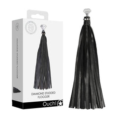 A black, diamond-studded whip with long leather straps is displayed next to its packaging. The package features an image of the whip and includes text reading "OUCH! Diamond Studded Whip" at the bottom, accompanied by a star symbol.