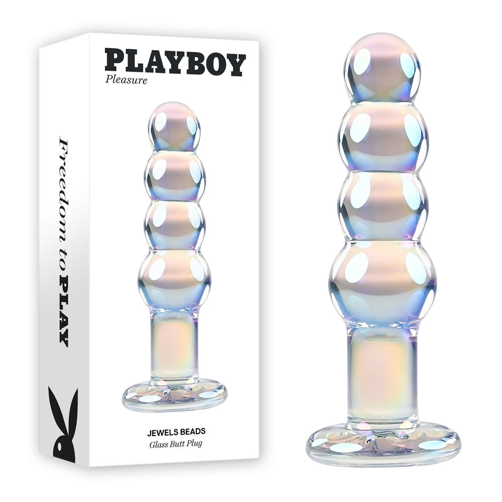 Image of a glass butt plug next to its packaging. The plug, crafted from borosilicate glass, features a series of round beads in ascending size and boasts an iridescent finish. The white packaging displays the text "PLAYBOY Pleasure JEWELS BEADS Glass Butt Plug.