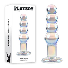 Image of a glass butt plug next to its packaging. The plug, crafted from borosilicate glass, features a series of round beads in ascending size and boasts an iridescent finish. The white packaging displays the text "PLAYBOY Pleasure JEWELS BEADS Glass Butt Plug.