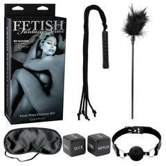 The Fetish Fantasy Series Limited Edition First Time Fantasy Kit includes a black whip, a feather tickler, a black eye mask, two black dice with words like "Kiss," "Suck," "Toes," and "Nipples," a black ball gag, and a box labeled "Fetish Fantasy Series" featuring an intimate couple—ideal for bondage beginners exploring their erotic playground.