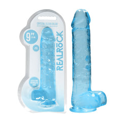 A clear blue REALROCK Realistic Dildo with Balls stands next to its transparent plastic packaging, which includes text stating "CRYSTAL CLEAR DILDO" and "REALROCK." The dildo is 9 inches in length, latex and phthalate free, with a flat suction base.