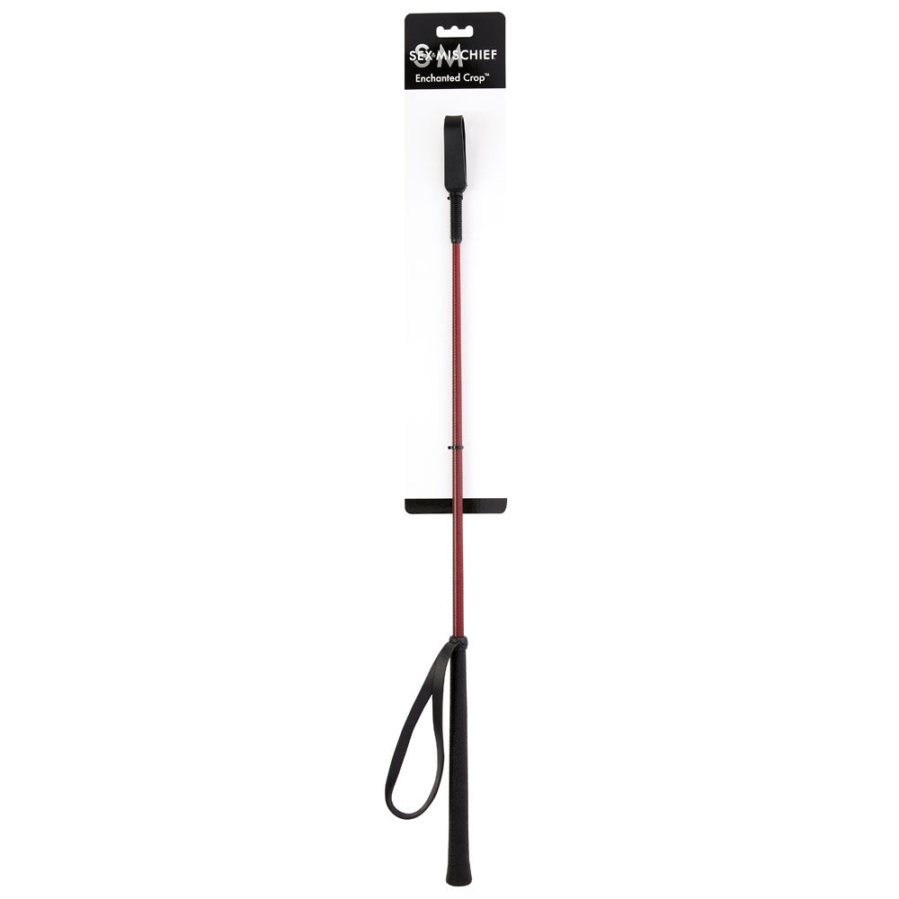 A red and black riding crop with a wrist strap is attached to a white packaging card labeled "Sex & Mischief Enchanted Crop." This carbon fibre crop features a black handle at one end and a small black loop at the other, making it an elegant impact toy for enthusiasts.