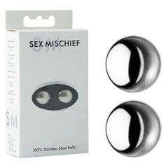 A box labeled "Sex & Mischief 100% Stainless Steel Balls" contains two stainless steel kegel weights. The front of the box features a small transparent window that reveals the balls inside. Two identical steel balls, perfect for kegel exercises to strengthen pelvic floor muscles, are also displayed outside of the box against a white background.