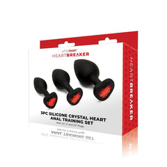A retail box labeled "WhipSmart Heartbreaker 3PC Silicone Crystal Heart Anal Training Butt Plug Set" contains three black silicone anal plugs in graduated sizes, each featuring a red heart-shaped crystal at the base. Perfect for body exploration, this set includes 2.5", 3", and 3.5" plugs.